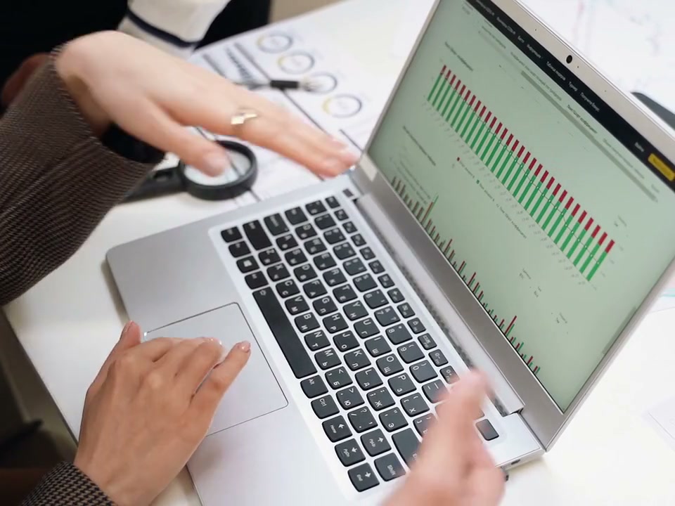 Bright overhead close-up of hands operating a laptop touchpad and gesturing at spreadsheet charts while reviewing printed documents; no faces.