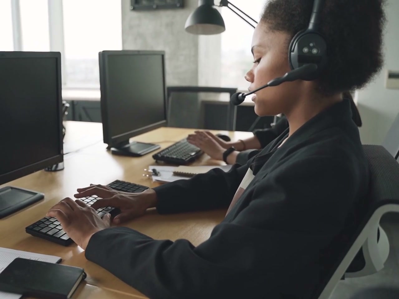 Side view of a headset support operator speaking while typing at a desktop workstation. Headset microphone, active hands and office equipment visible; dark monitor surfaces do not show a CRM interface.