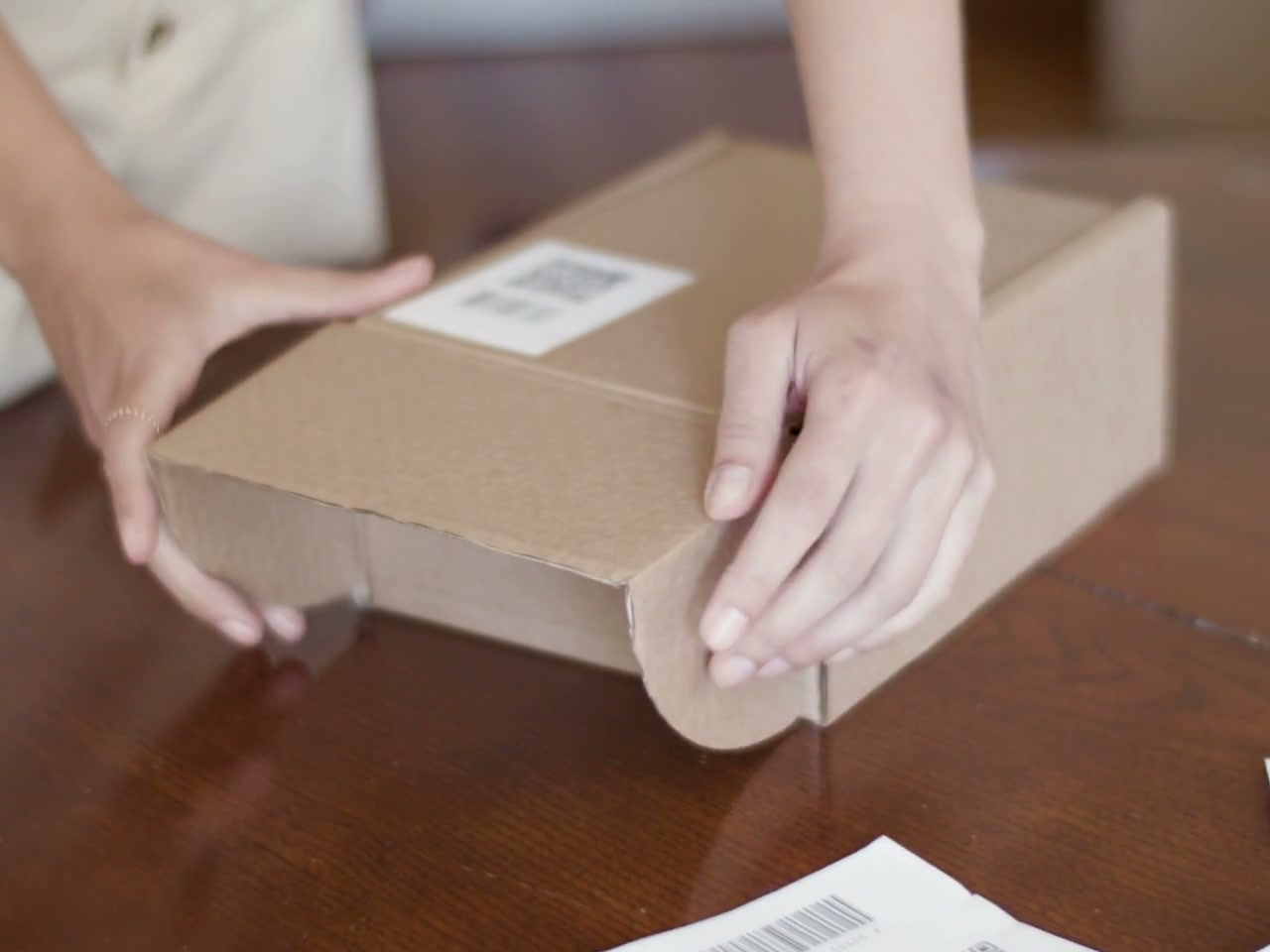 Hands close and rotate a cardboard parcel on a clean wooden packing table with shipping barcode labels; no faces.