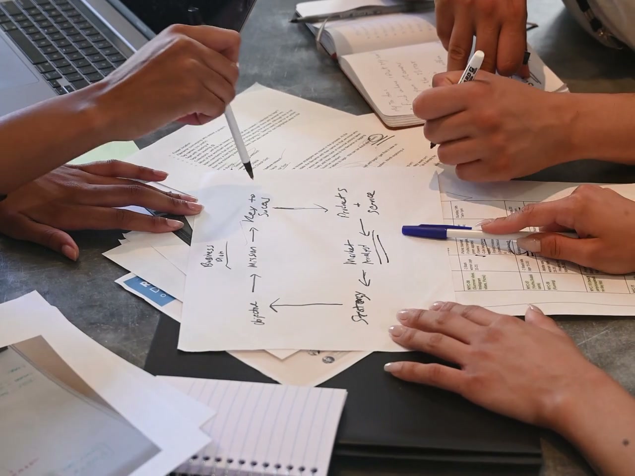 Several colleagues point at and mark a handwritten planning diagram on a gray meeting table, surrounded by printed documents, notebooks and laptops. Faces are excluded. General project-planning scene; no specific merchandise is shown.