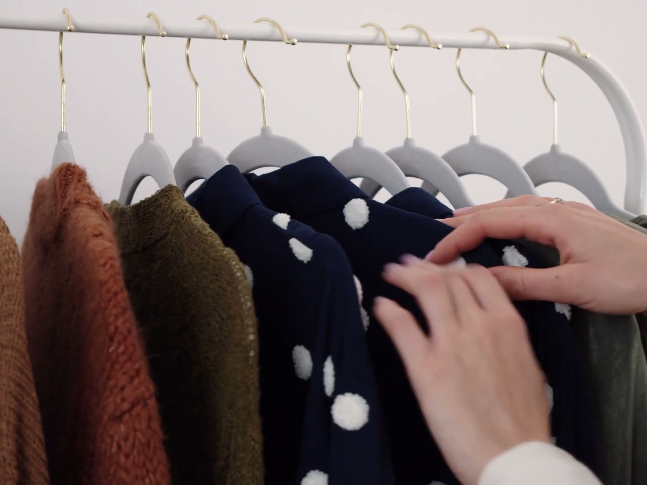 Close-up hands inspect and select knit garments on a white clothing rail; no faces. Neutral cream, brown and navy products.