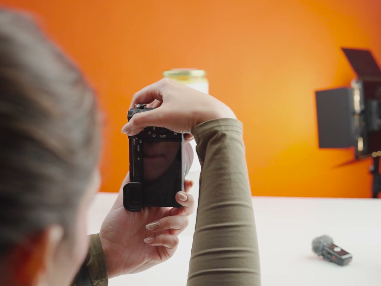 Over-the-shoulder close-up of hands using a mirrorless camera to photograph a bottle on a studio table; orange sweep and dedicated studio light visible. Camera screen activates during the clip.